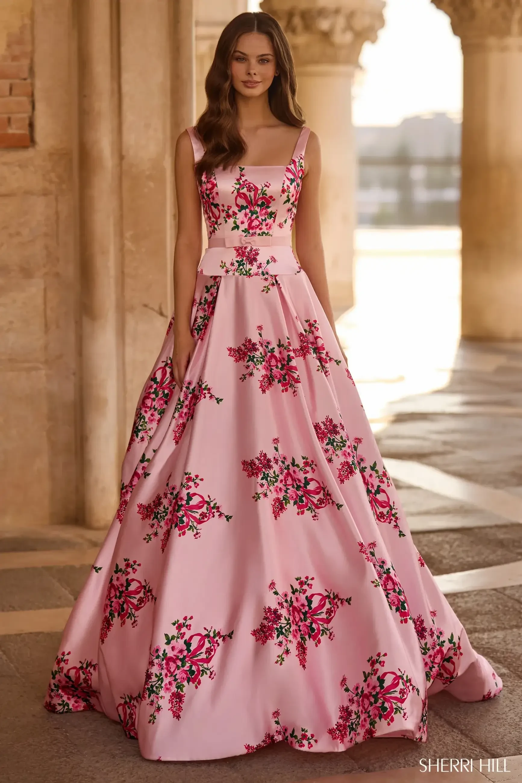 A woman in a flowing pink gown adorned with vibrant floral patterns stands elegantly in a sunlit, arched stone corridor. The scene feels romantic and regal.