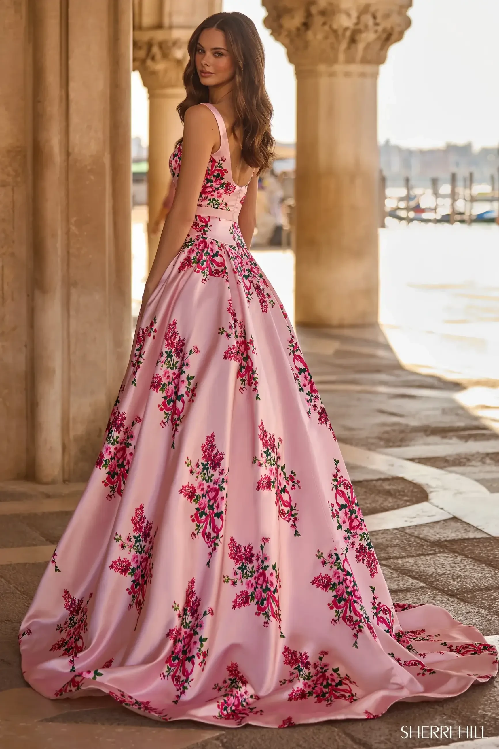 A woman in a flowing pink gown adorned with vibrant floral patterns stands elegantly in a sunlit, arched stone corridor. The scene feels romantic and regal.