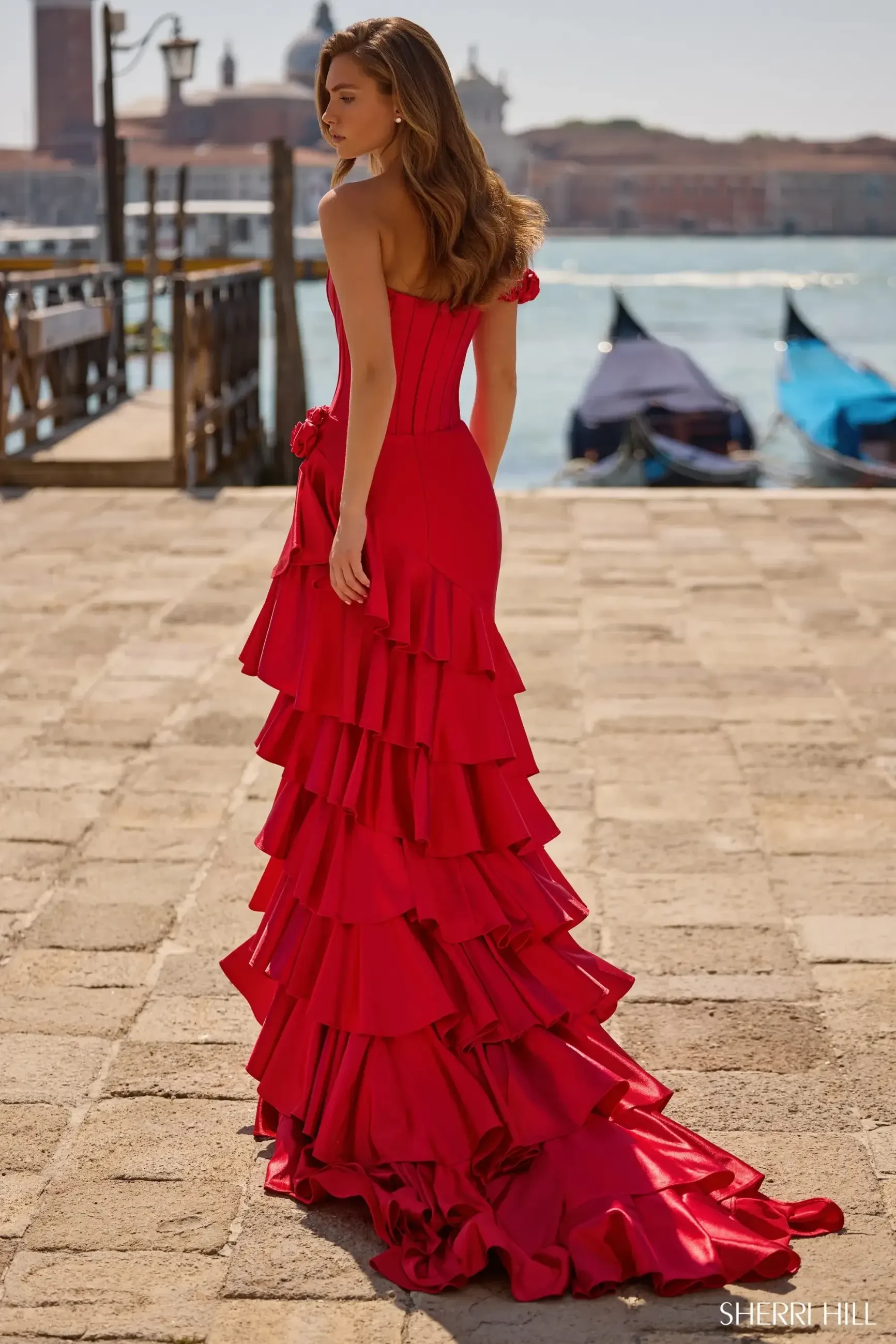 A woman in a vibrant red, off-shoulder gown with ruffled layers stands elegantly on a sunlit stone path by a shimmering waterfront, exuding glamour.